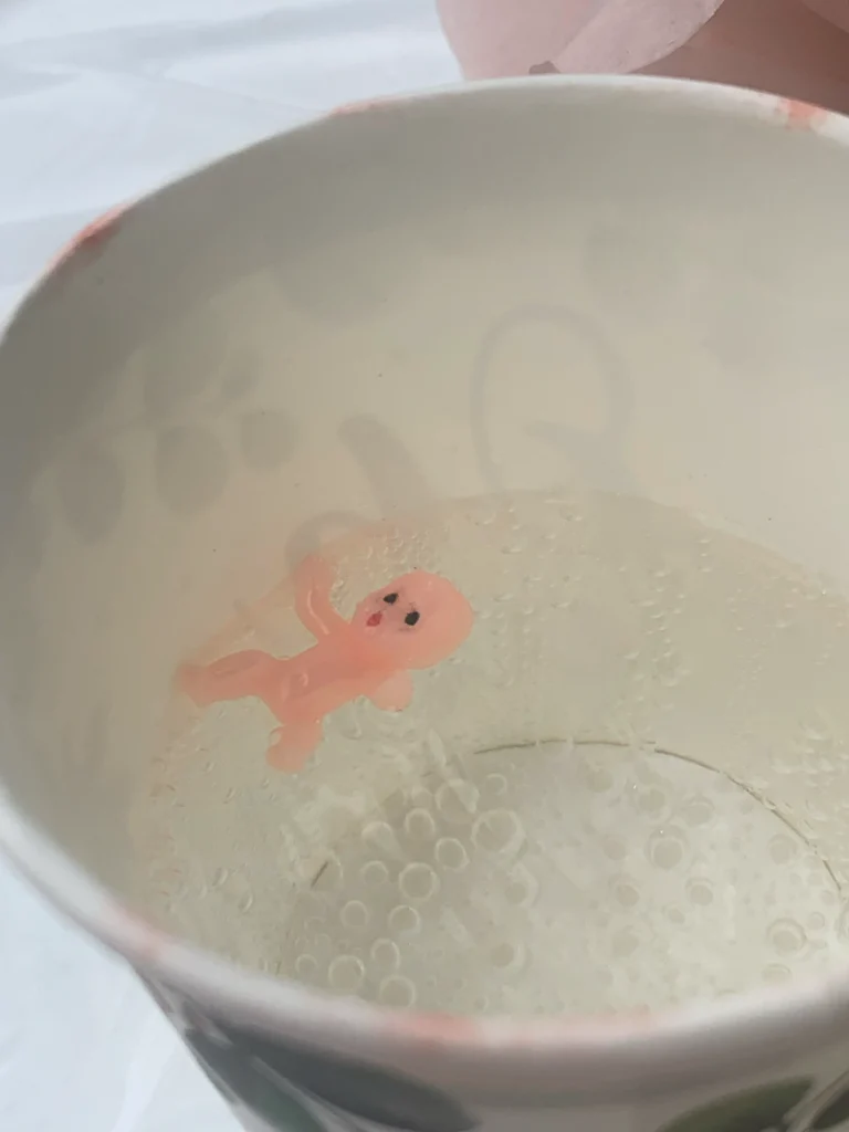 A King Cake plastic baby figurine floats in a cup of bubbly water.